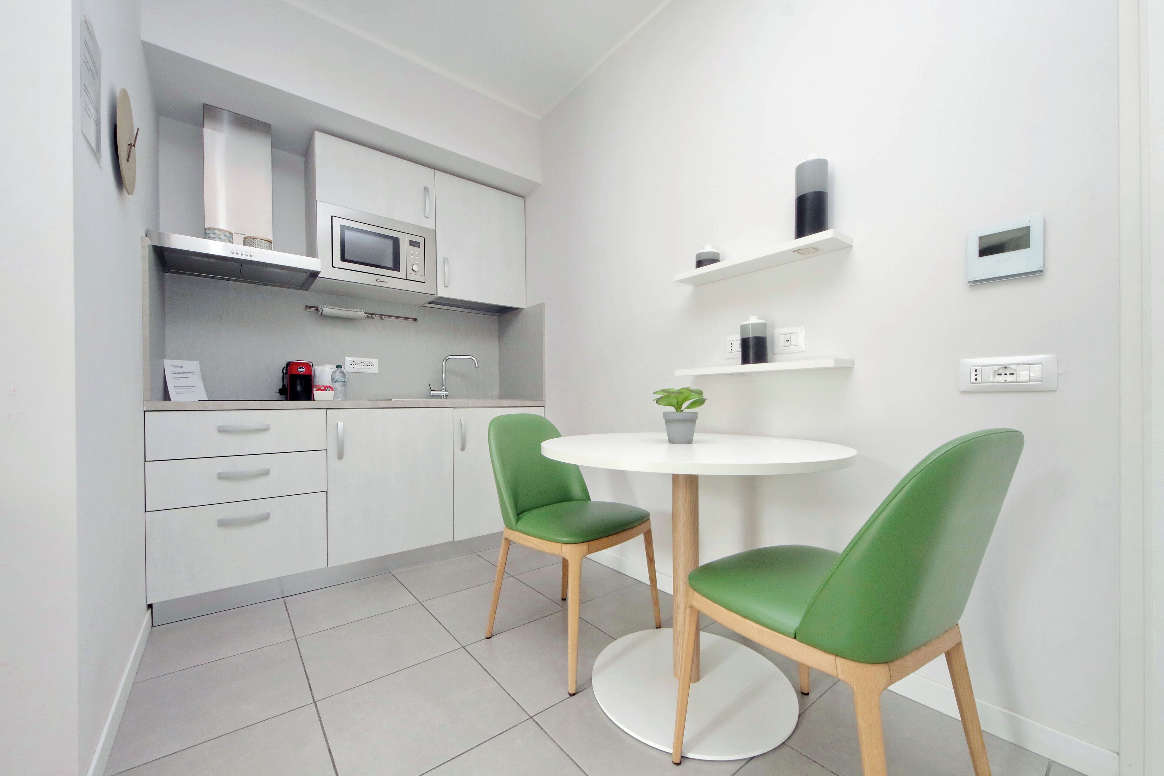 A modern kitchen with white cabinets, a microwave, and a coffee machine. There is a round white table with two green chairs and a potted plant.