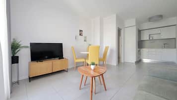 Ein modernes Wohnzimmer mit einem Flachbildfernseher, einem Holzschrank, einem runden Holztisch mit einem Topf und einer Pflanze, gelben Stühlen und einem grauen Sofa.