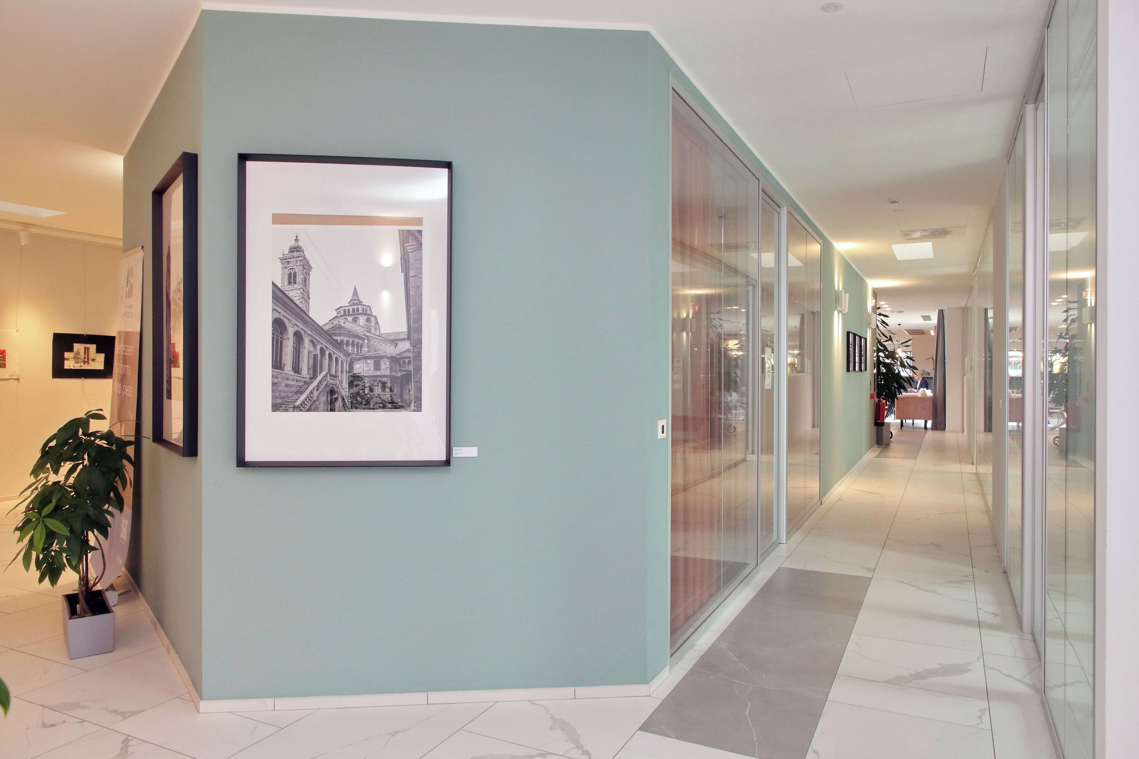 A corridor with framed pictures on the wall, a potted plant, and a glass partition.