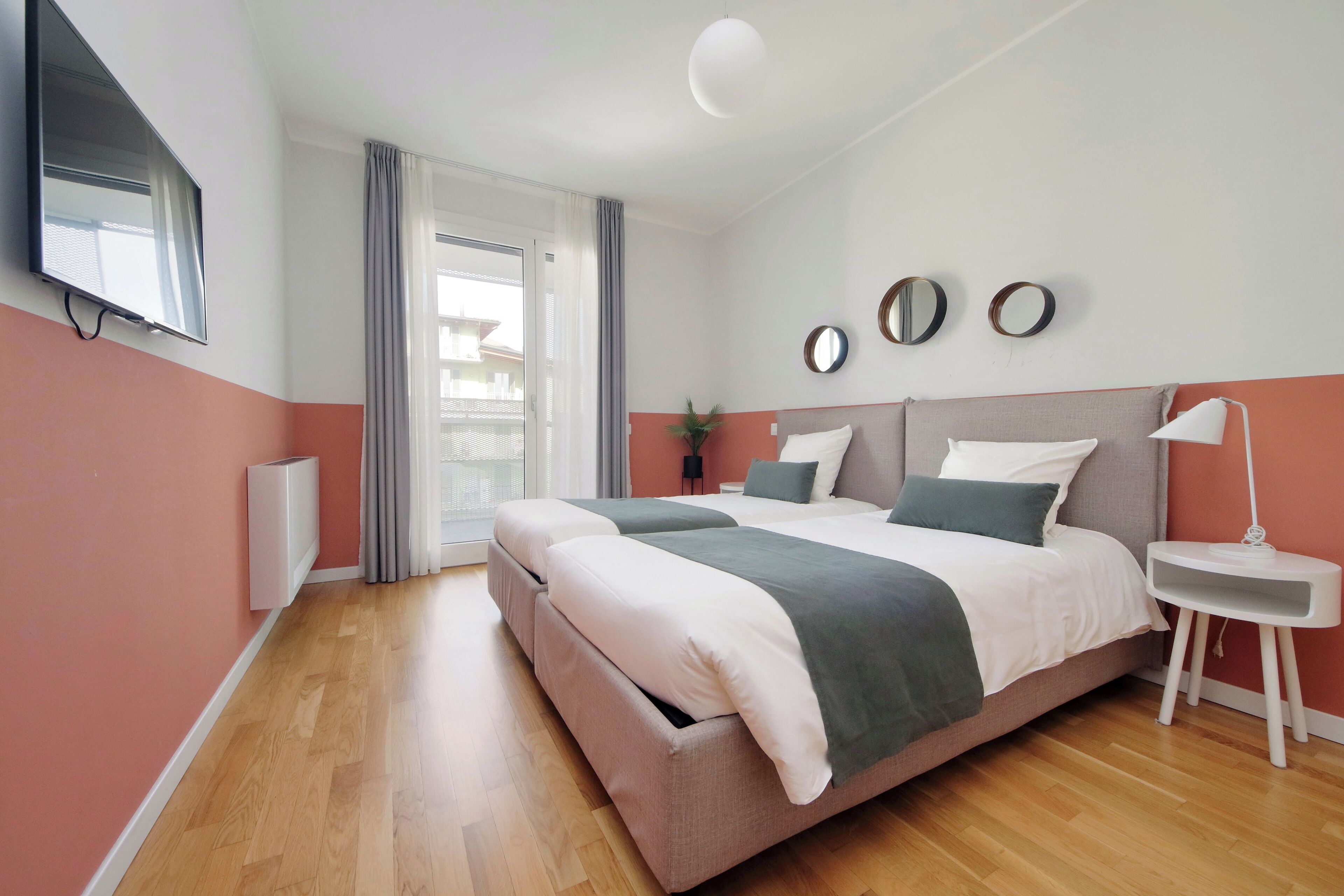 A modern bedroom with a large bed, a bedside table, a TV mounted on the wall, and a sliding glass door leading to a balcony.