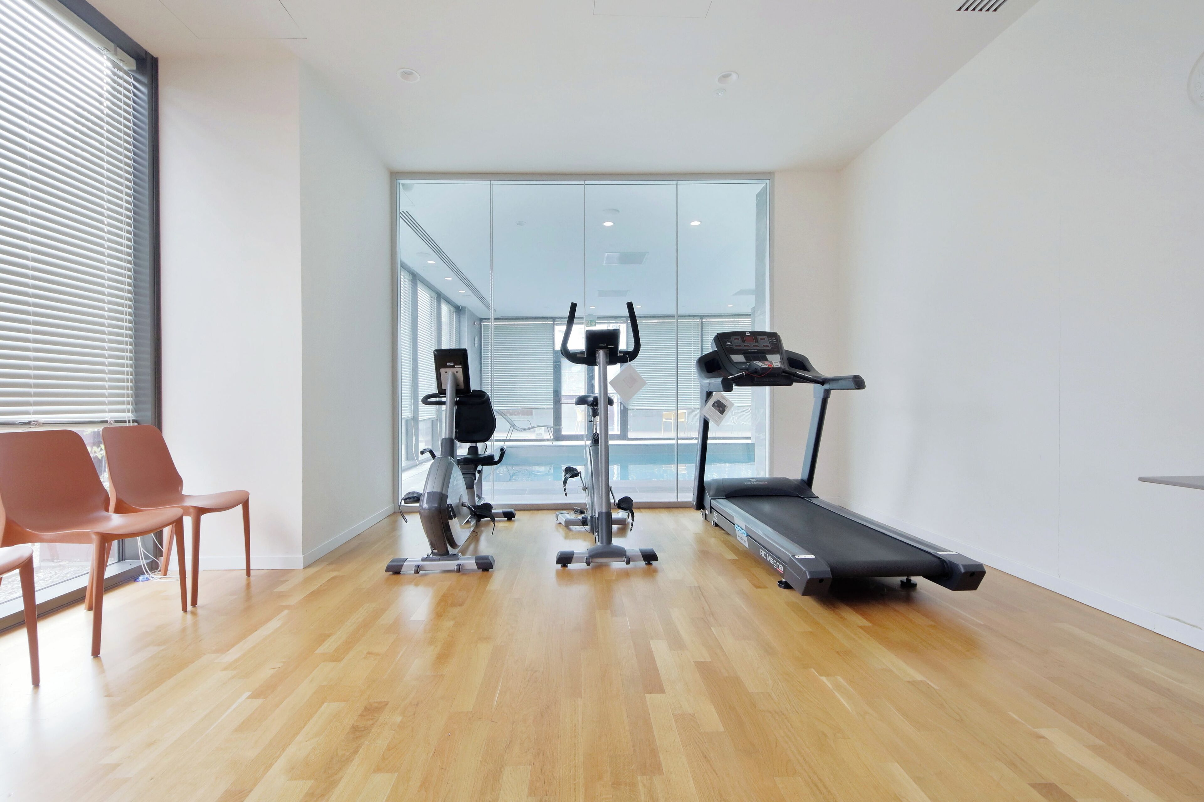 A modern gym with a treadmill, elliptical, and weight machine, wooden flooring, and large windows.
