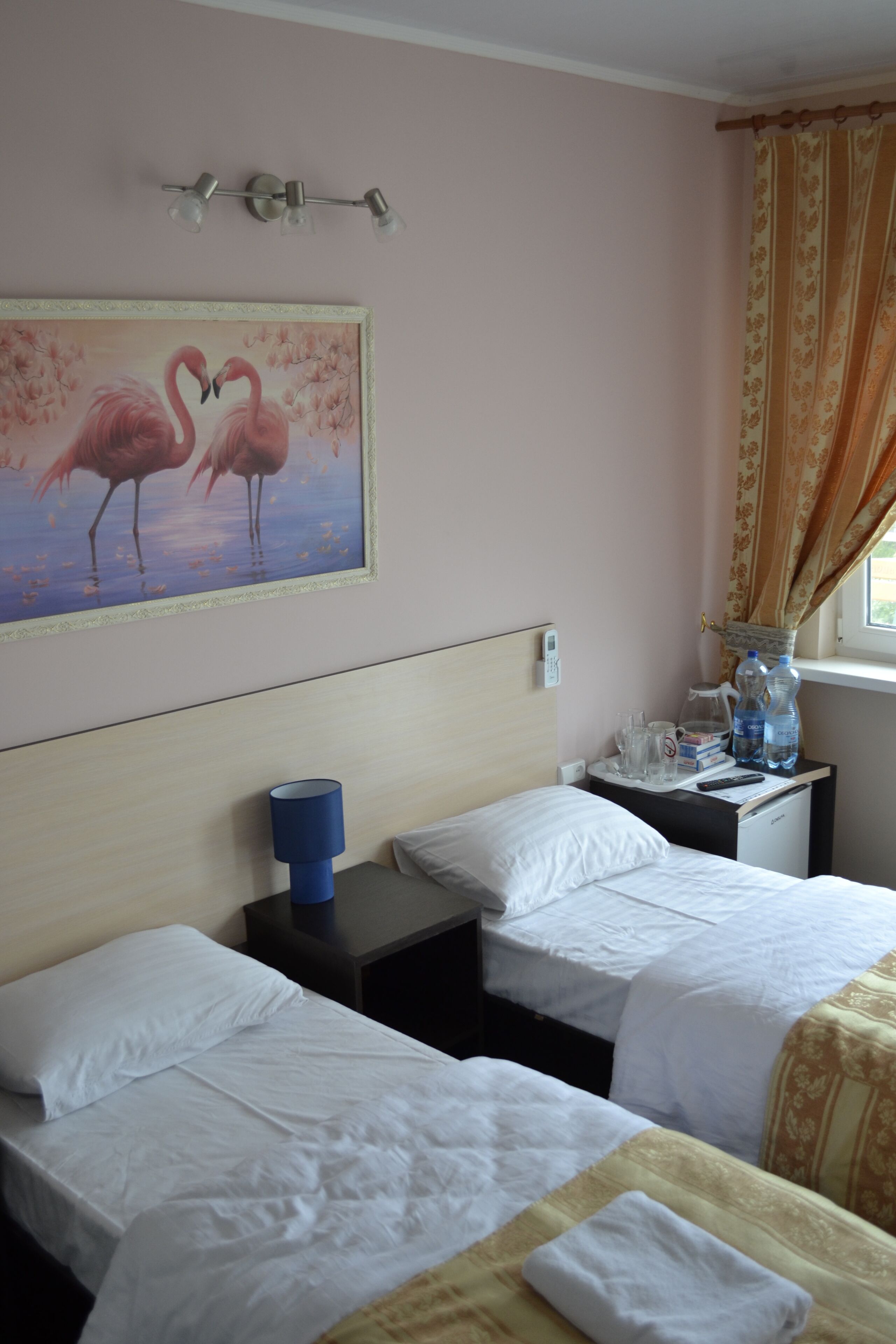 Ein Hotelzimmer mit zwei Betten, einem Flamingo-Gemälde, einem Nachttisch mit Lampe und einem fenster mit Vorhängen.