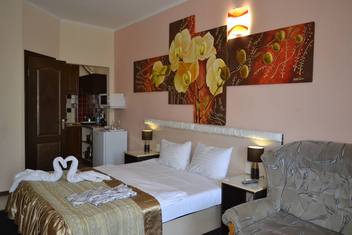 A hotel room with a bed, a sofa, a painting of flowers on the wall, and a kitchenette in the background.