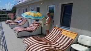 A sunlit outdoor area with striped lounge chairs and a poolside table, a person standing by the pool, and a building with a number '32' sign. - Shellman (Odesa)
