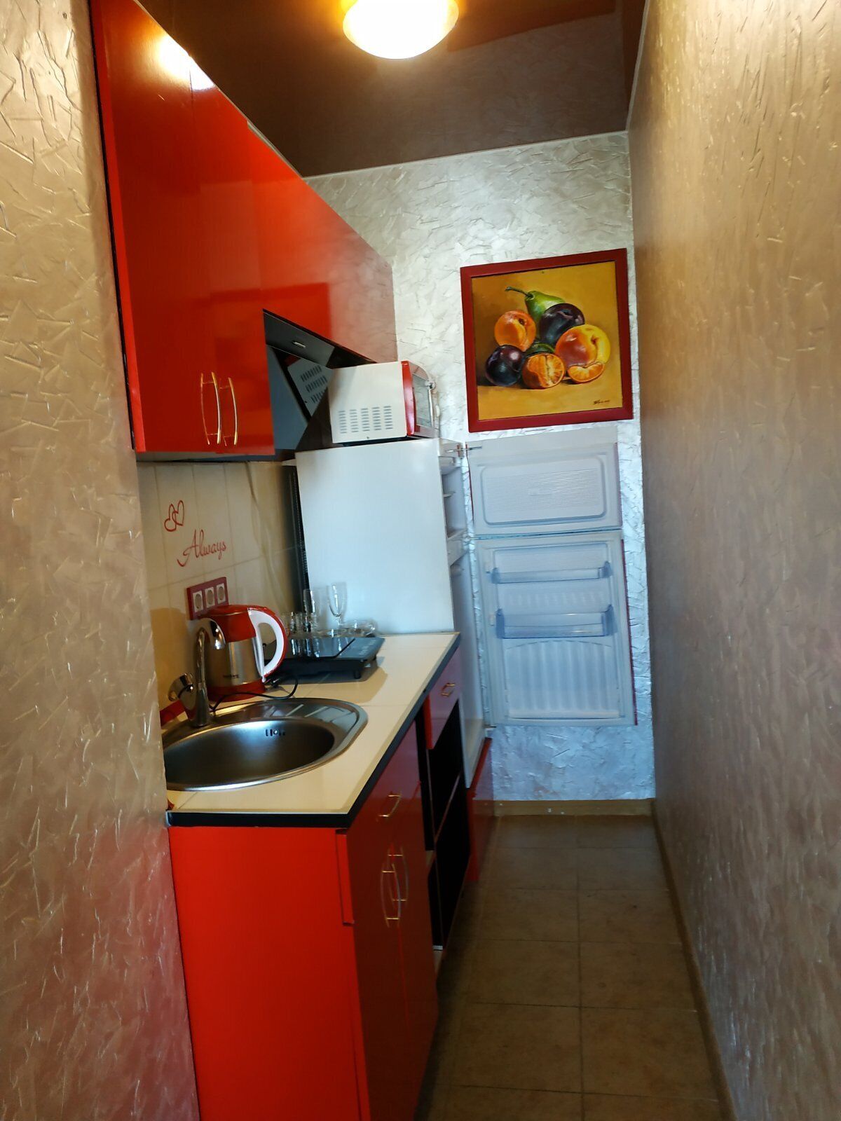 A compact kitchen with red cabinets, a white refrigerator, a microwave, and a sink. There is a framed picture of fruits on the wall.