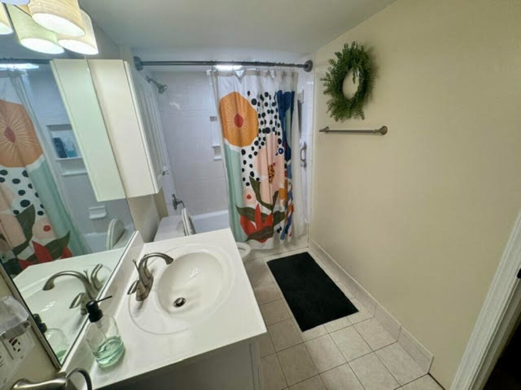 Un baño con un lavabo blanco, un espejo, una cortina de ducha con estampado floral colorido y un felpudo negro en el suelo.