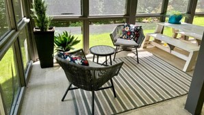 A sunlit patio with wicker chairs and a table, large windows, and a view of greenery.