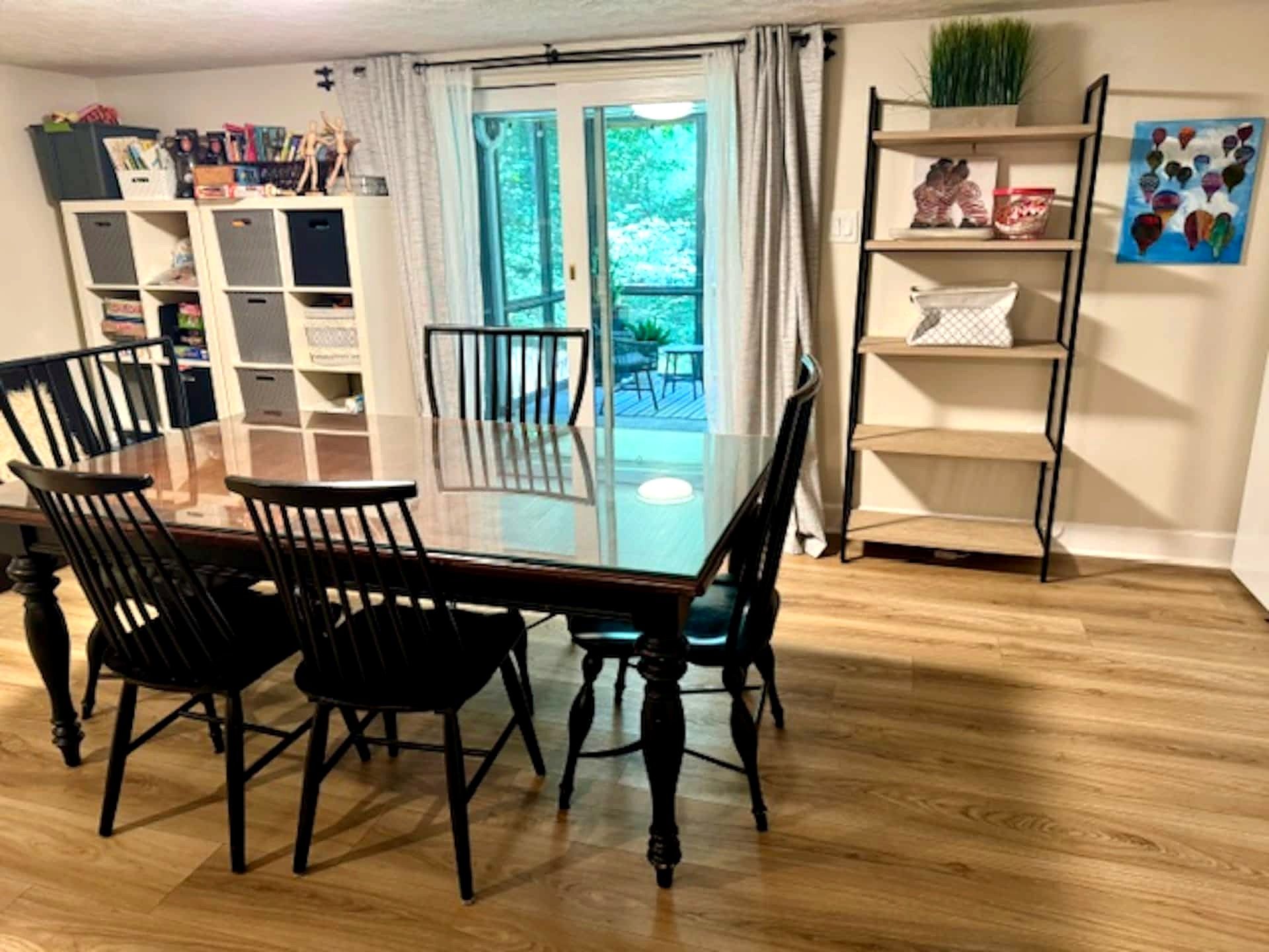 A dining area with a glass-top table and chairs, a bookshelf, and a view through a glass door to an outdoor deck.