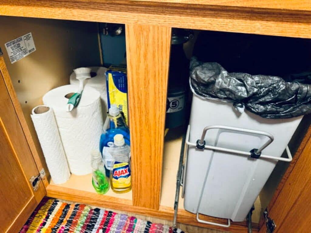 Una kitchenette pequeña con un refrigerador, un cubo de basura, una botella de detergente para platos, un rollo de toallas de papel y una lata de café.