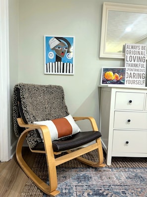 A room with a rocking chair, a white dresser, and a framed artwork on the wall.