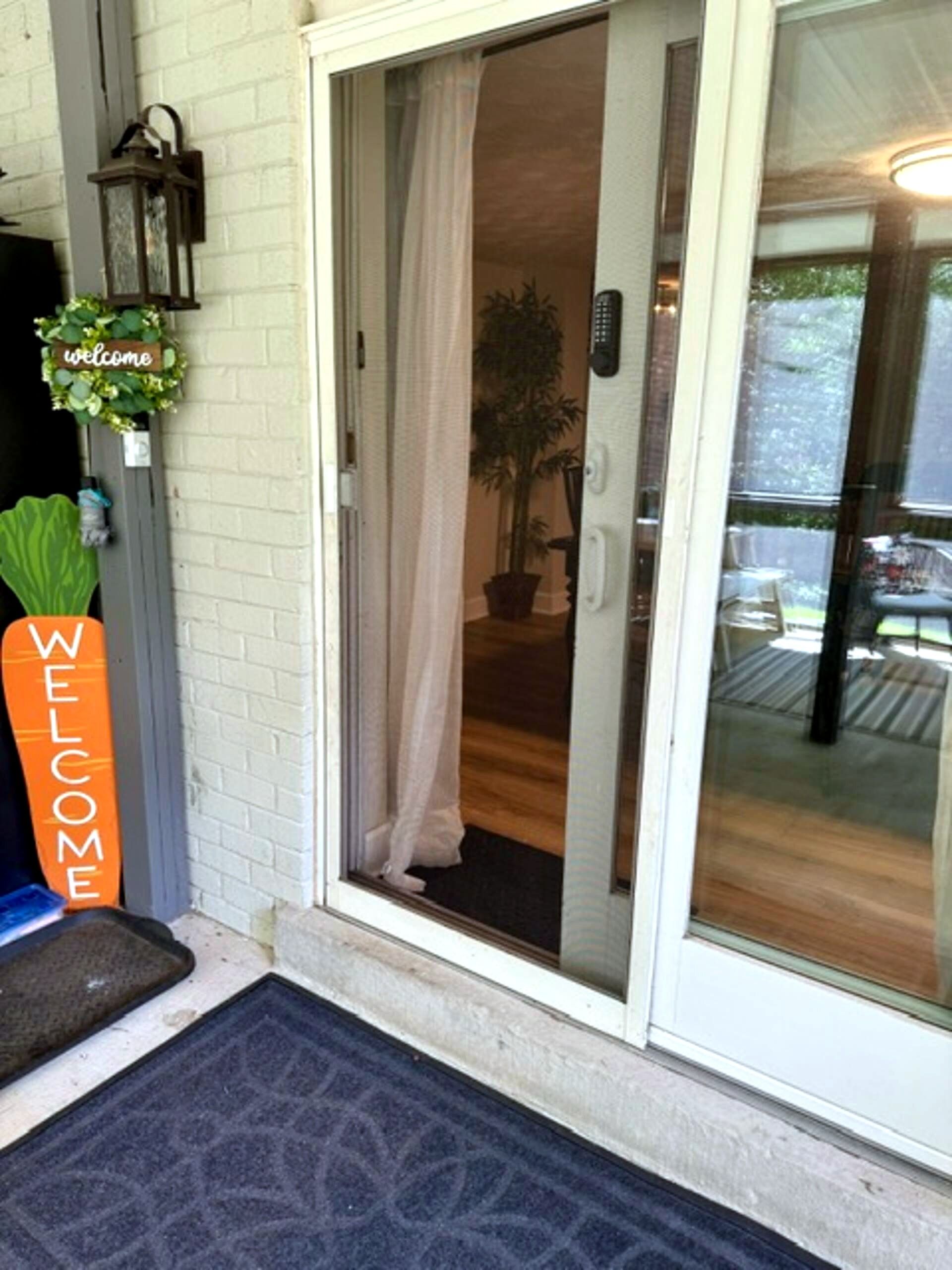 A sliding glass door entrance with a welcome sign, a potted plant, and a remote control on the wall.