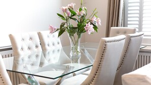 Ein Esstisch mit Glasplatte, vier gepolsterten Stühlen, einer Vase mit rosa Blumen und einem Fenster mit Jalousien im Hintergrund.