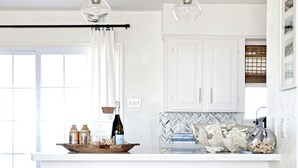 A kitchen with white cabinets, a herringbone backsplash, two bar stools, and pendant lights. - Salt Air Cottage: Beachfront Charming & Luxe home! (Galveston)