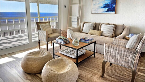 A cozy living room with a view of the ocean, featuring wicker furniture, a coffee table with books, and a painting on the wall. - Salt Air Cottage: Beachfront Charming & Luxe home! (Galveston)