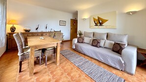Una sala de estar con una mesa de comedor de madera, sillas de mimbre, un sofá gris con cojines estampados, una alfombra y un cuadro en la pared.