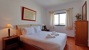 Una cama bien hecha con sábanas blancas, una lámpara de noche, un cuadro en la pared, una ventana con cortinas y un armario de madera.