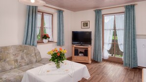 Un soggiorno accogliente con un divano, una TV su un mobile in legno, un tavolino con un vaso di fiori e ampie finestre con tende.