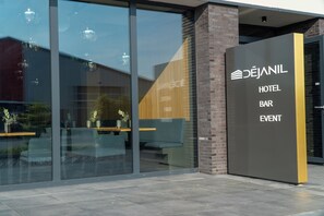 Entrada de un hotel con fachada de vidrio, zona de estar y cartel que indica servicios de hotel, bar y eventos.