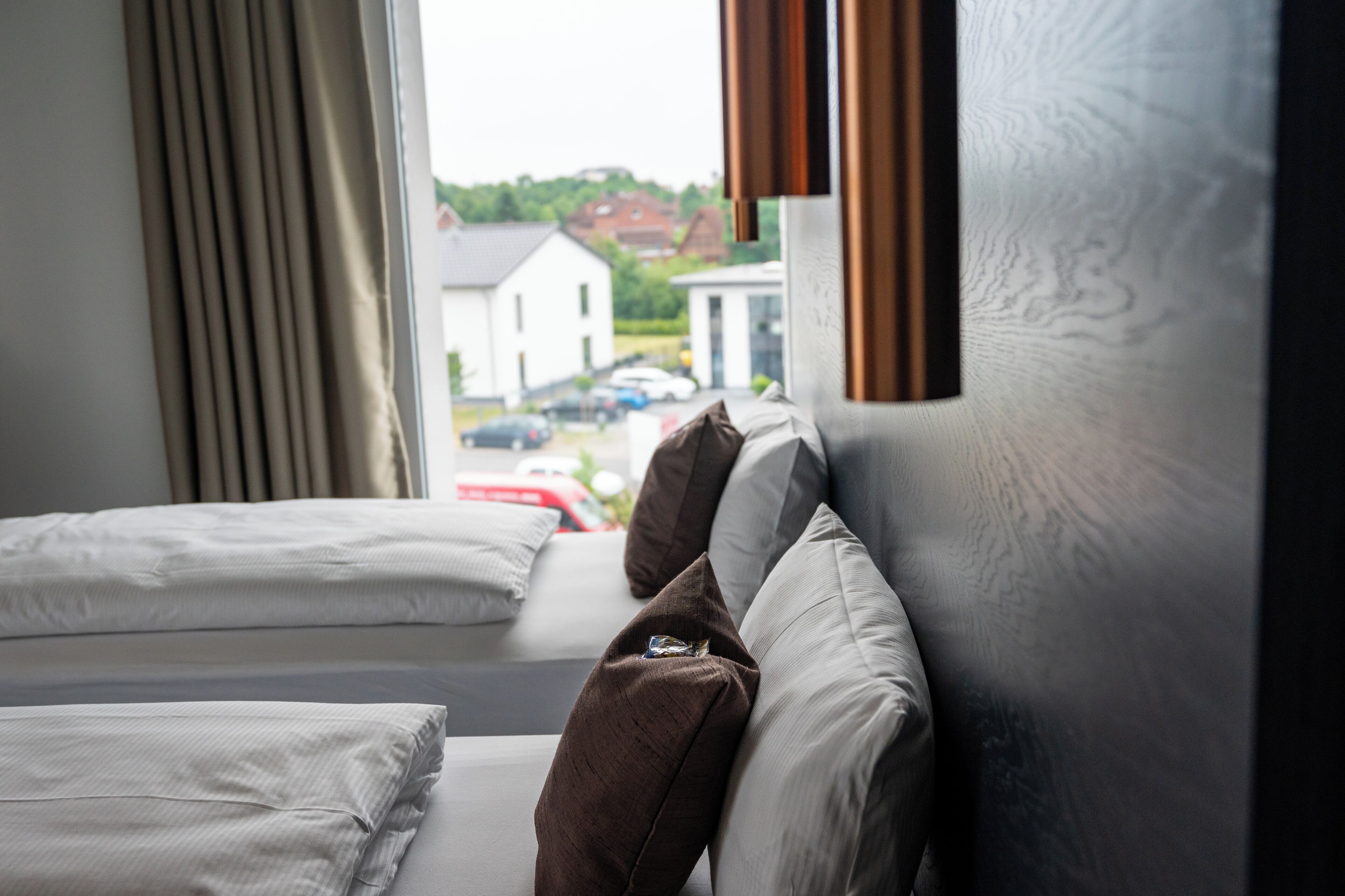 Een hotelkamer met twee bedden, een raam met gordijnen en uitzicht op een straat met geparkeerde auto's en een gebouw.