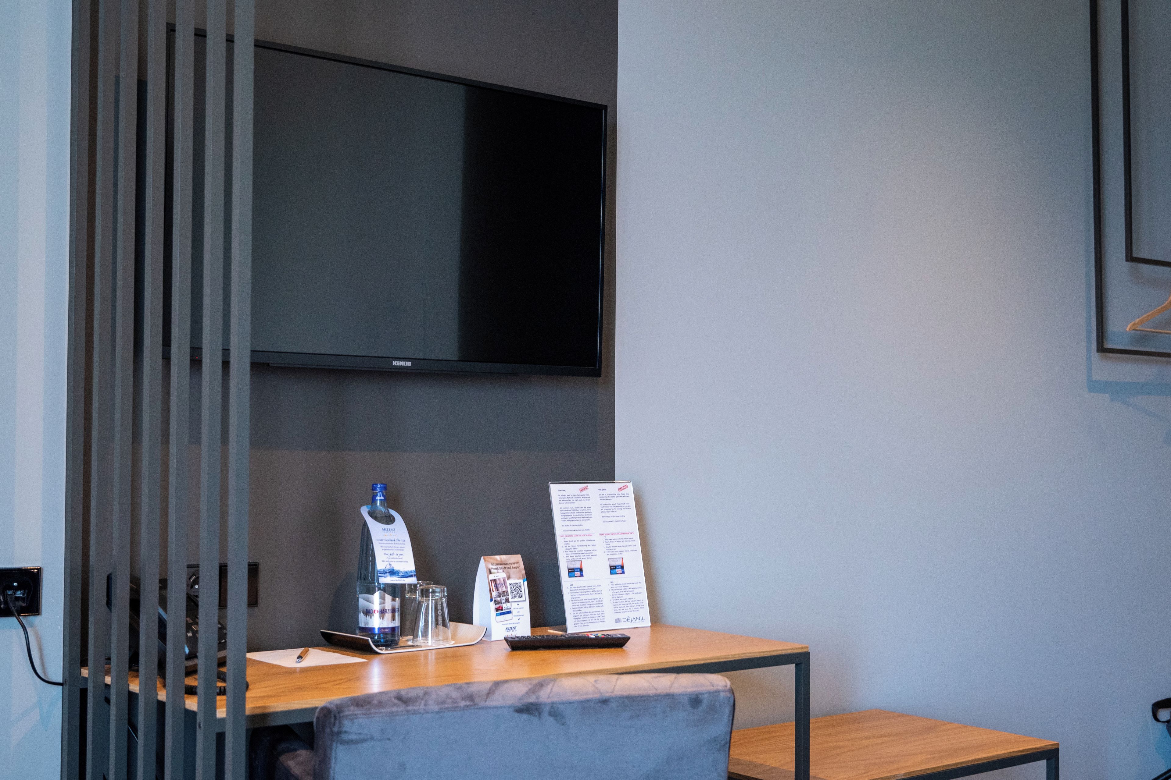 A hotel room with a flat-screen TV mounted on the wall, a wooden desk with a remote control, a glass bottle, and a brochure.