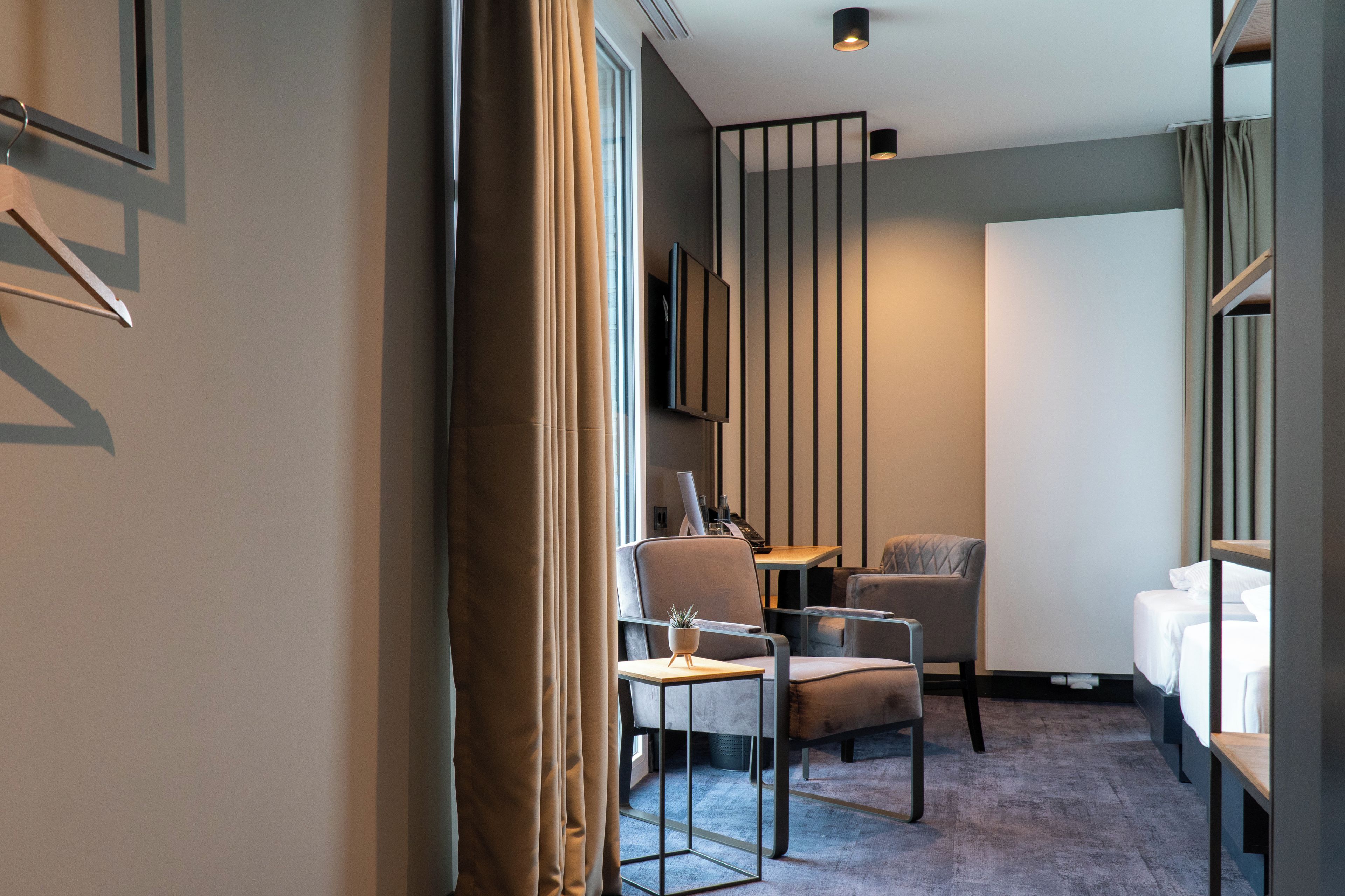 A modern hotel room with a bed, two chairs, a desk, and a large window.