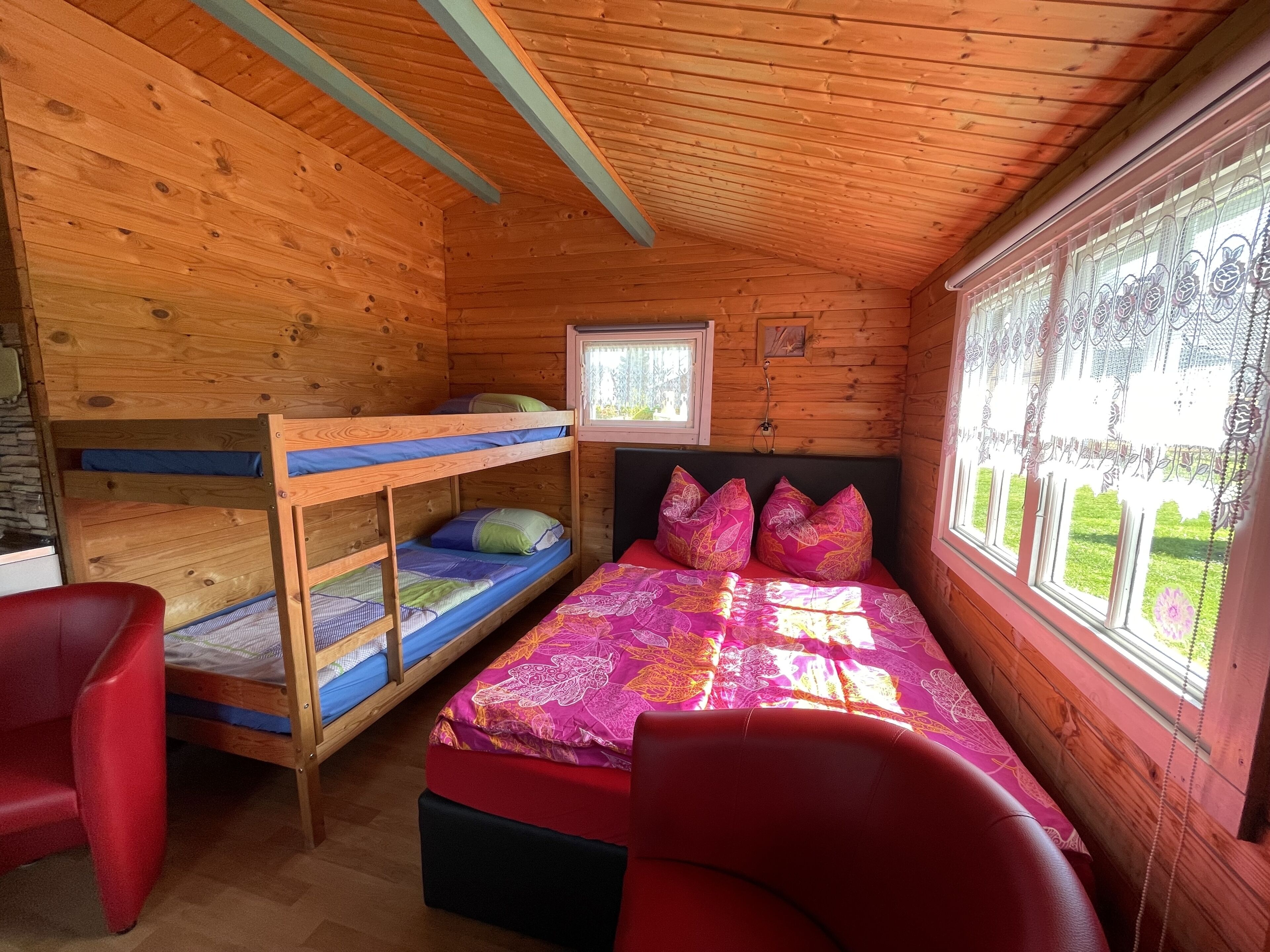 Ein Holzhüttenraum mit Etagenbetten, einem Einzelbett mit farbenfroher Bettdecke, einem roten Sessel und einem Fenster mit Spitzgittervorhängen.
