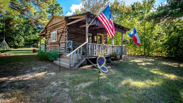 A rustic log cabin with a porch, an American flag, and a French flag. - Log Cabin - TINY HOUSE on acreage (Montgomery)