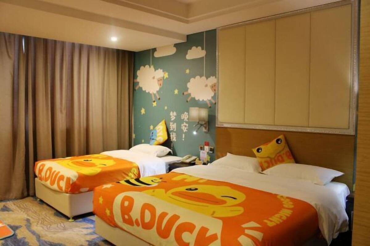 Habitación de hotel con dos camas, un ventanal grande con cortinas, una pared decorada con nubes y ovejas, y una colcha con diseño de patos.