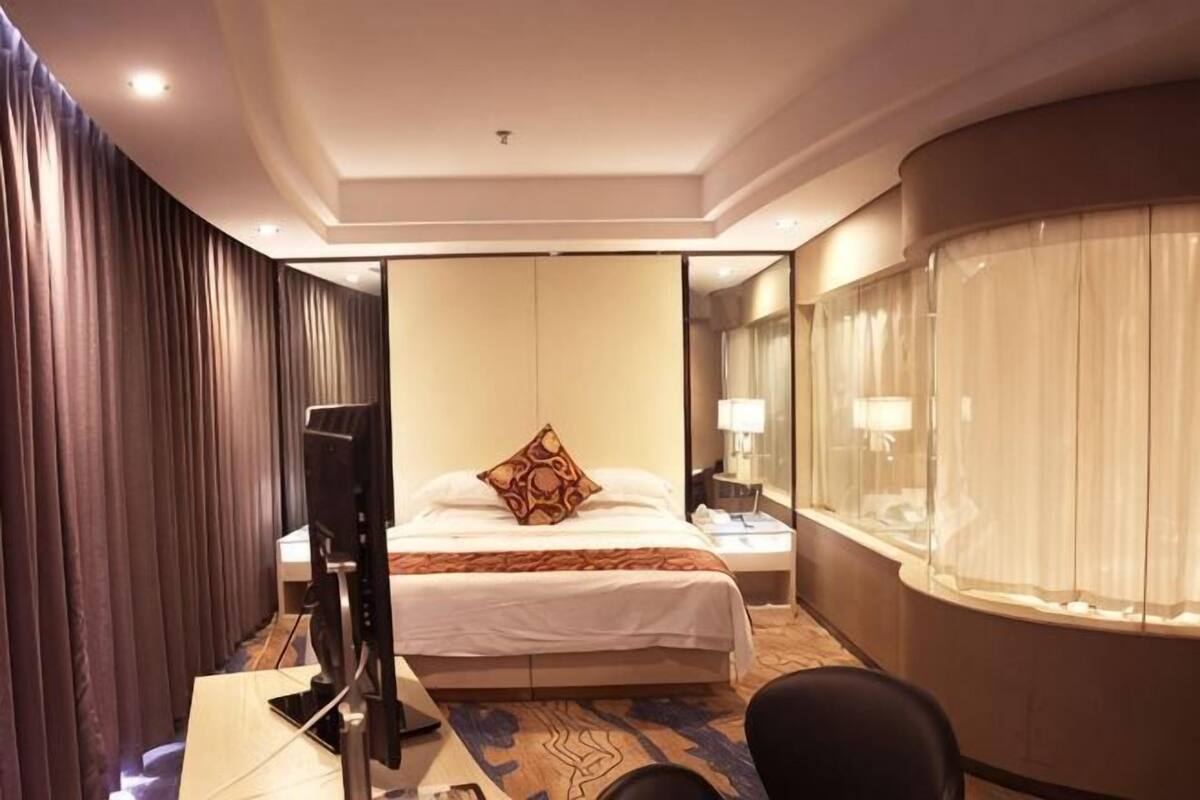 Habitación de hotel con una cama, un escritorio con ordenador, televisor, lámpara y ventana con cortinas.