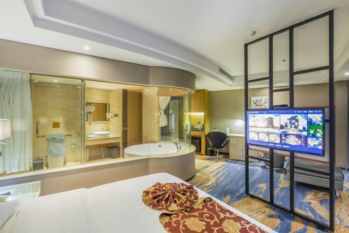 Habitación de hotel con baño de vidrio, una cama grande, televisor de pantalla plana y piso alfombrado.