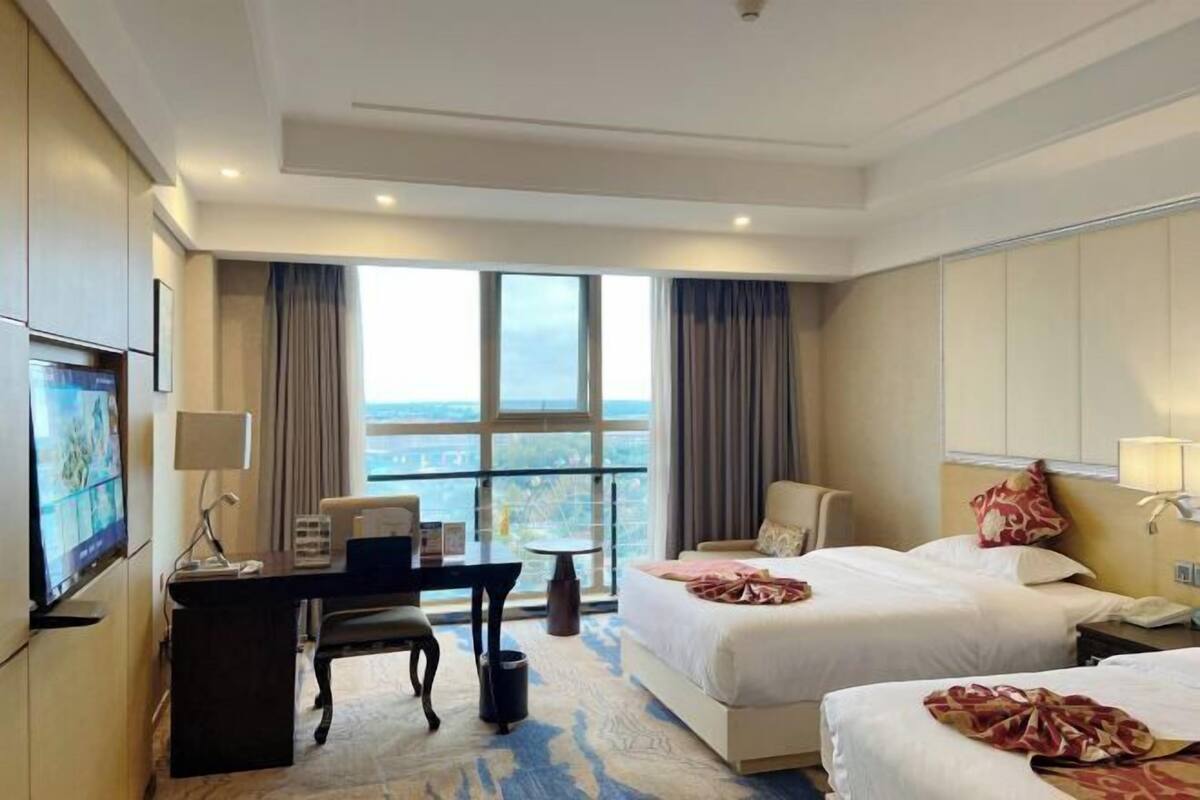 Habitación de hotel con dos camas, un escritorio, una silla, un televisor y un ventanal grande con vistas a la ciudad.