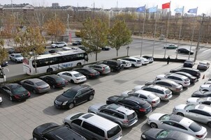 Un estacionamiento con varios autos y un autobús, rodeado de árboles y banderas.