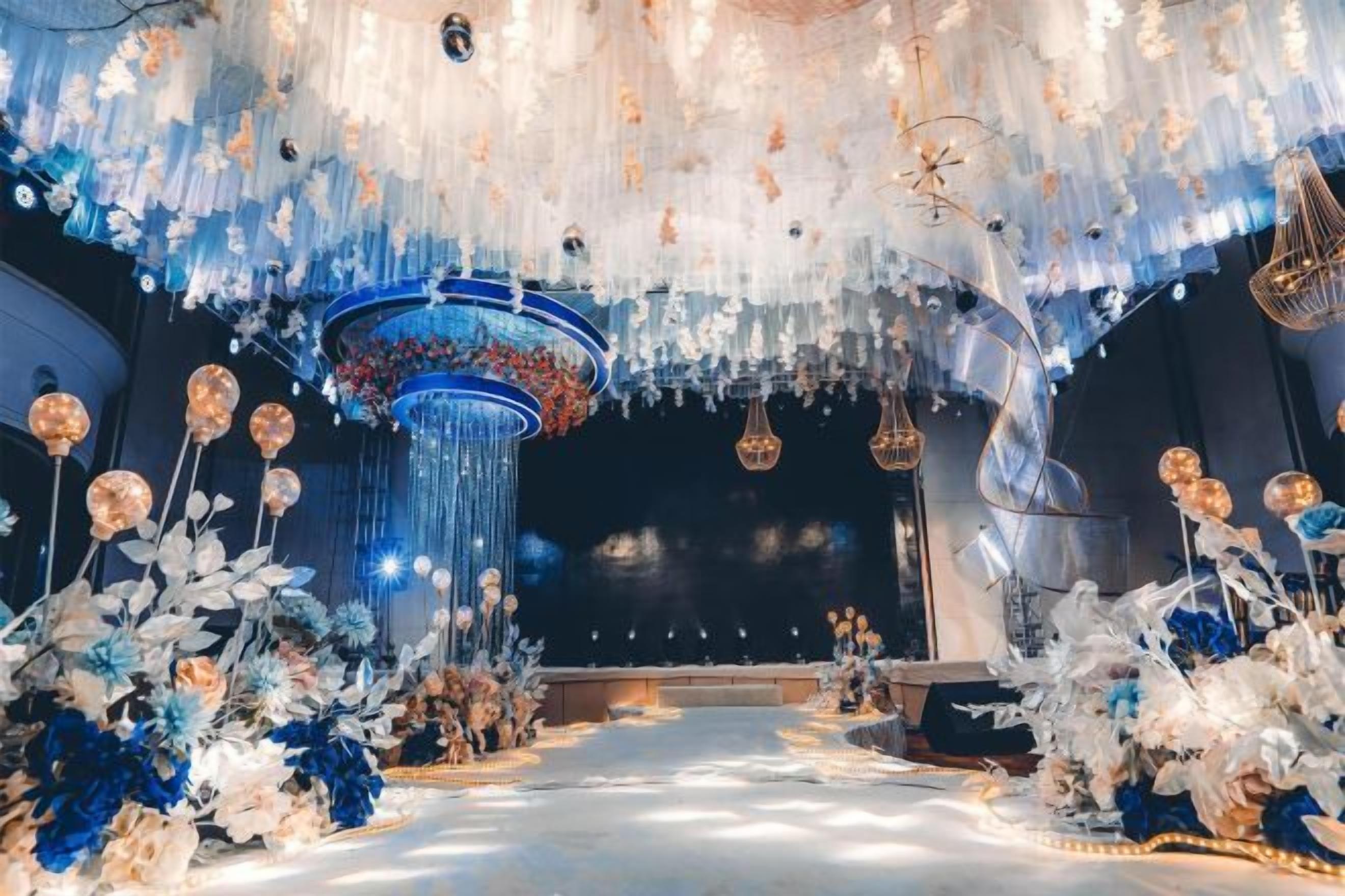 Un espacio interior con un escenario central, elementos decorativos colgantes y arreglos florales.