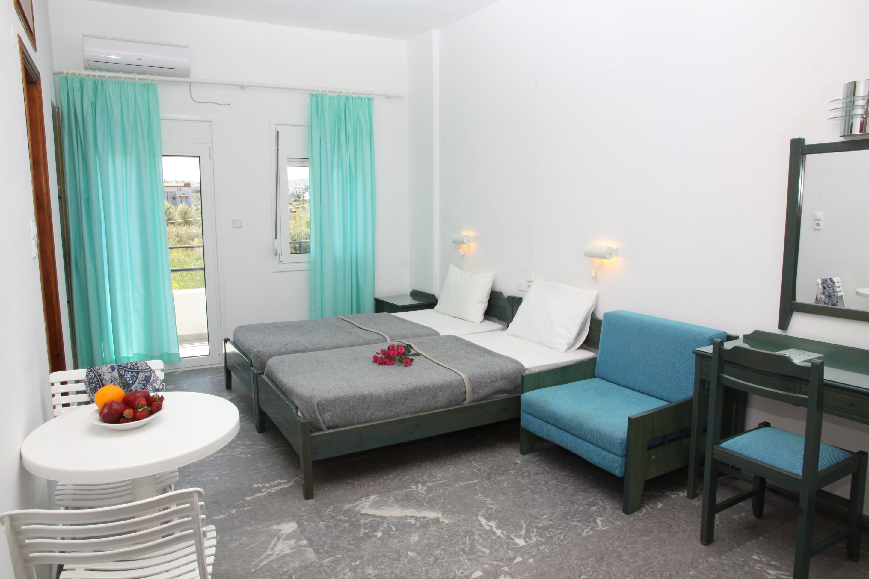 A bedroom with two beds, a desk, a chair, a small table with fruit, and a window with curtains.
