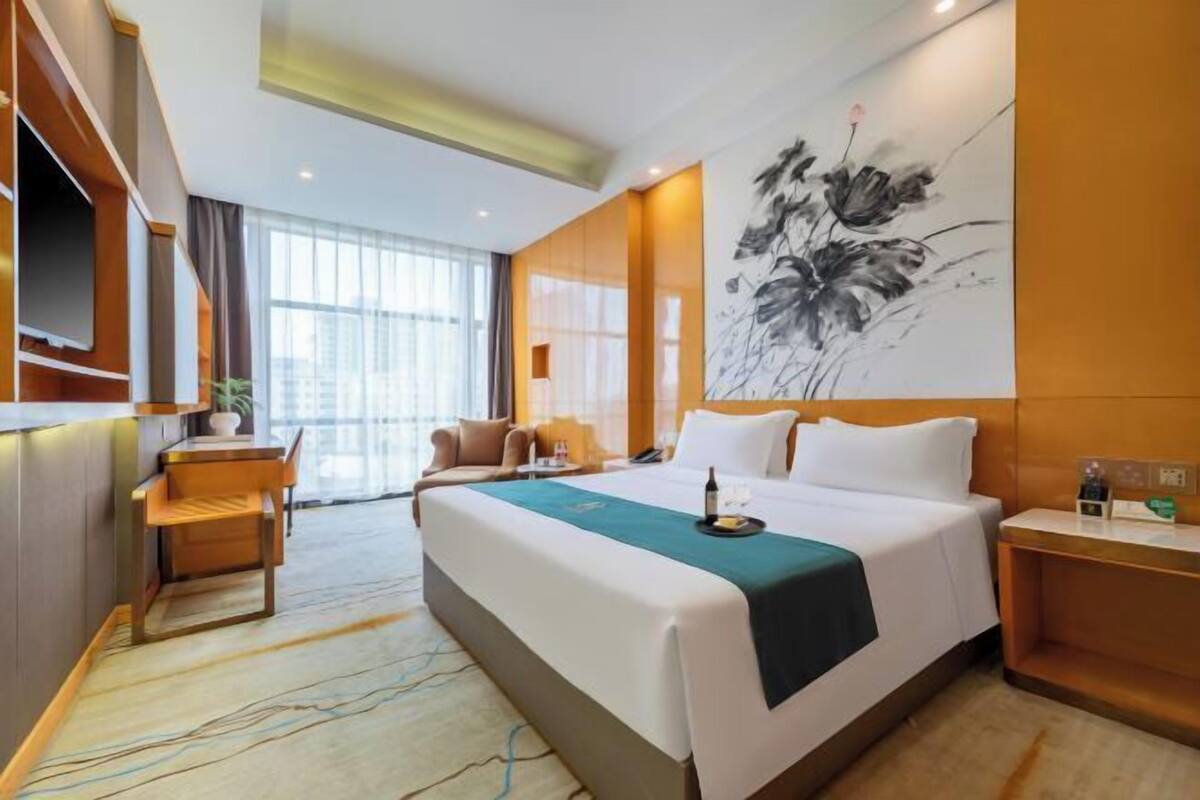 A hotel room with a bed, a desk, a chair, a large artwork on the wall, and a window with curtains.
