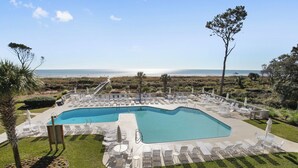A pool area with lounge chairs and umbrellas, surrounded by greenery and a view of the ocean.