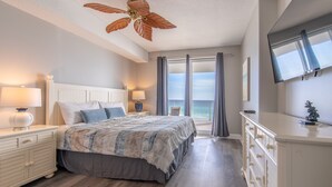 Ein Schlafzimmer mit einem Bett, Nachttischen, einer Kommode, einem Deckenventilator, einem großen Fenster mit Vorhängen und einem an der Wand montierten Fernseher.
