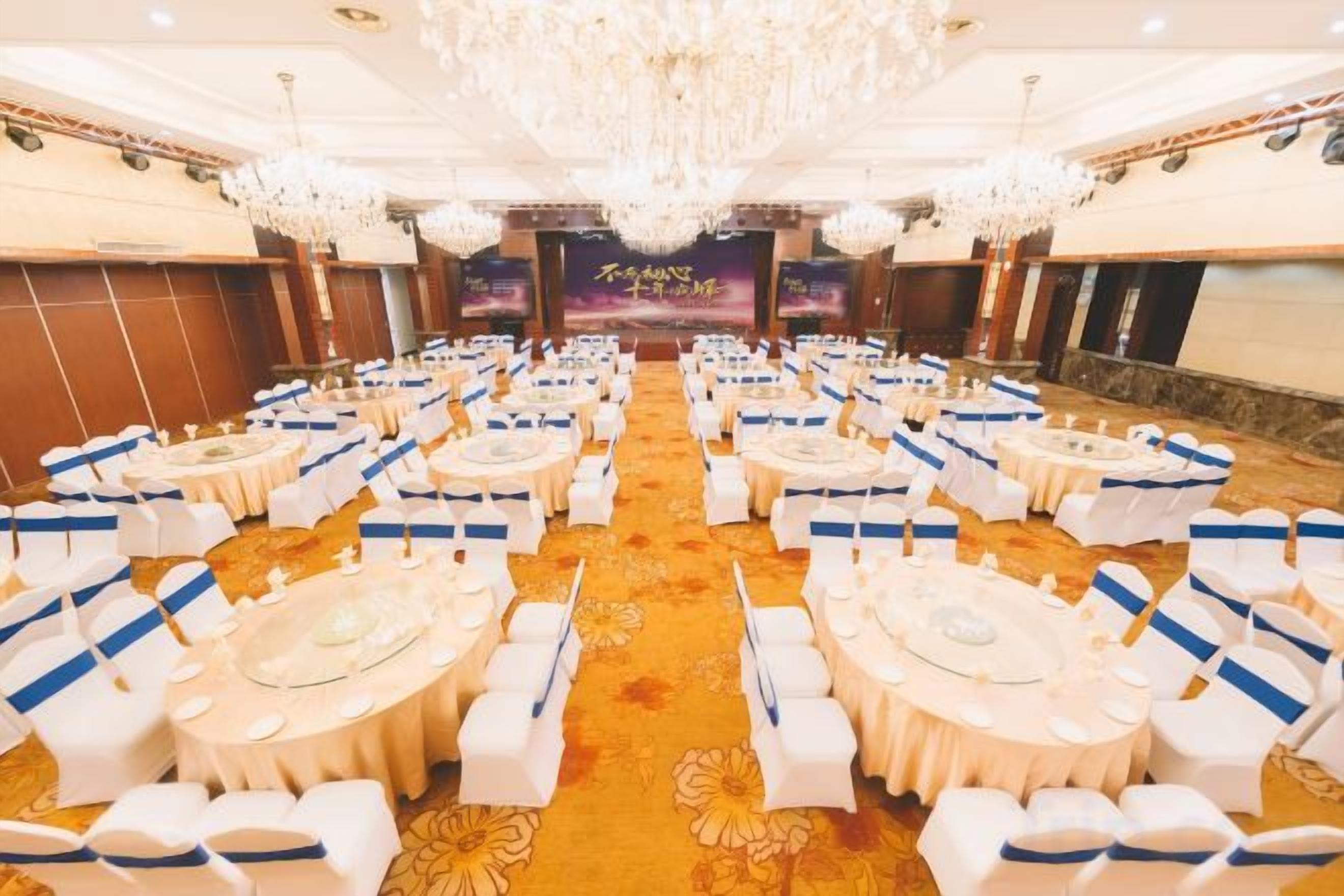 A banquet hall with round tables and chairs, a chandelier, and a stage with a banner.