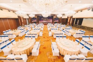 Un salón de banquetes con mesas y sillas redondas, una araña y un escenario con un estandarte.