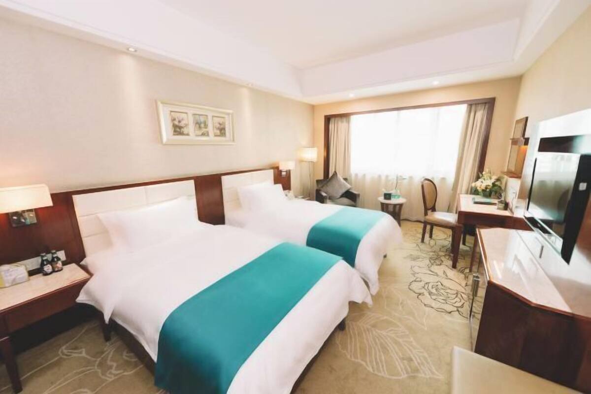 Ein Hotelzimmer mit zwei Betten, einem Schreibtisch, einem Sessel, einem Fernseher und einem Fenster mit Vorhängen.