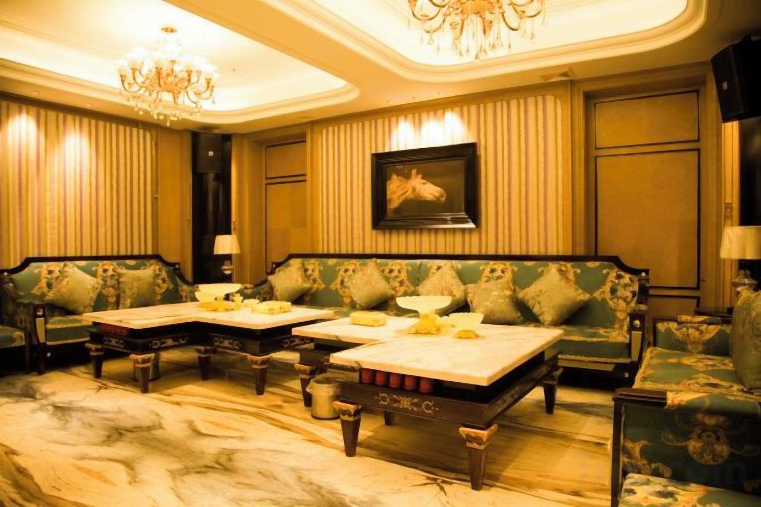 A room with a chandelier, green upholstered sofas, a marble floor, and a framed picture on the wall.