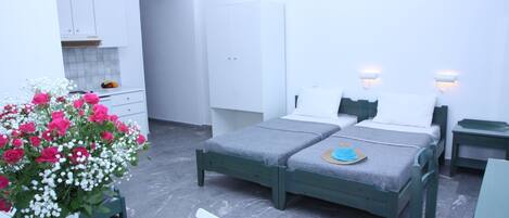 A bedroom with two beds, a small table, a chair, a vase of flowers, and a kitchenette in the background.