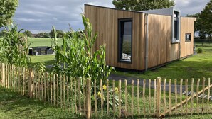 Une cabane en bois moderne, dotée d’une grande fenêtre et d’une porte, entourée d’une clôture en bois et d’une zone herbeuse.