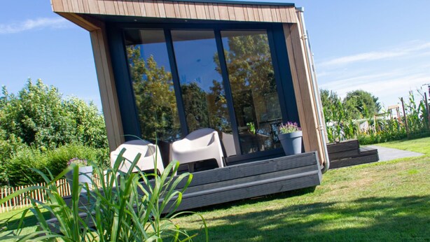 Une maison moderne, de plain-pied, dotée de grandes fenêtres en verre, d’un toit plat et d’une petite terrasse avec des meubles d’extérieur.