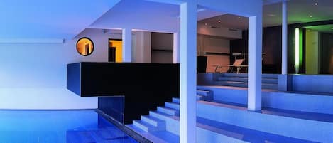 A modern indoor swimming pool with a glass floor, a staircase, and a circular window.