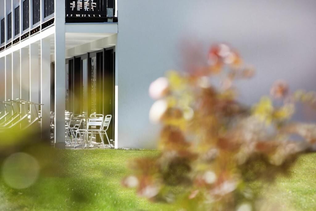 Un edificio moderno con un letrero en idioma extranjero, un área de estar al aire libre techada y un primer plano borroso con vegetación.