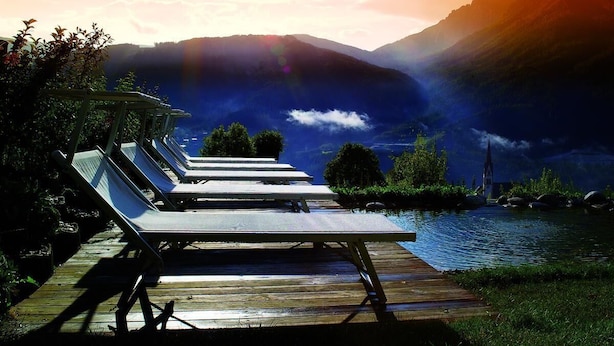 Um deck de madeira com espreguiçadeiras com vista para um lago, rodeado por montanhas e uma igreja.
