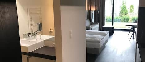 Quarto de hotel moderno com uma cama grande, espaço de estar separado e vista para o exterior.