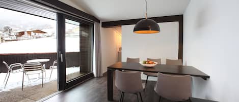 Un comedor moderno con una mesa de madera oscura, cuatro sillas y una lámpara colgante grande. Una puerta corredera de cristal da a un patio exterior con sillas y vistas a un paisaje nevado.