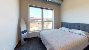 Une chambre d’hôtel avec une grande fenêtre, un lit doté d’une tête de lit grise et une planche de surf appuyée contre le mur.